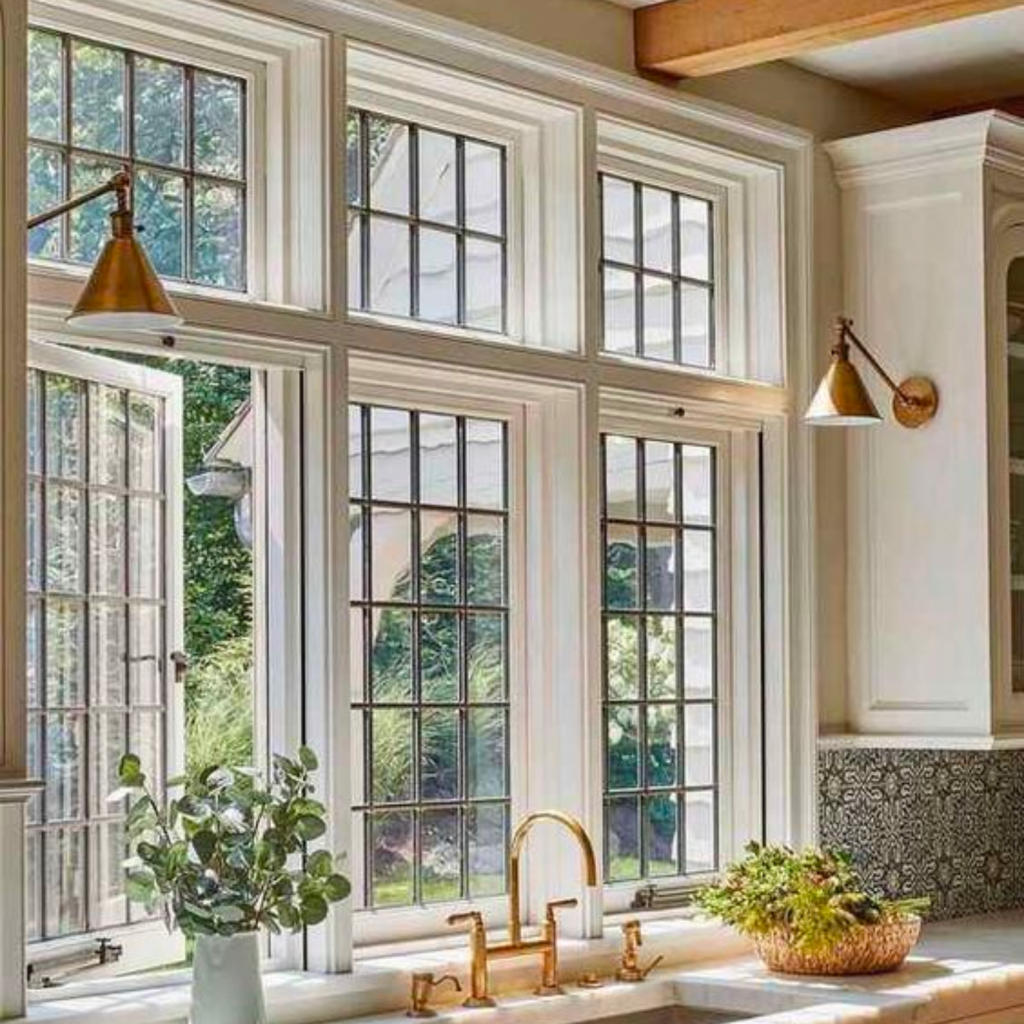 Beautiful tall kitchen frame windows, above a sink.  One of the three windows is ajar.   There are two flanking sconces on either side of the window opening. 