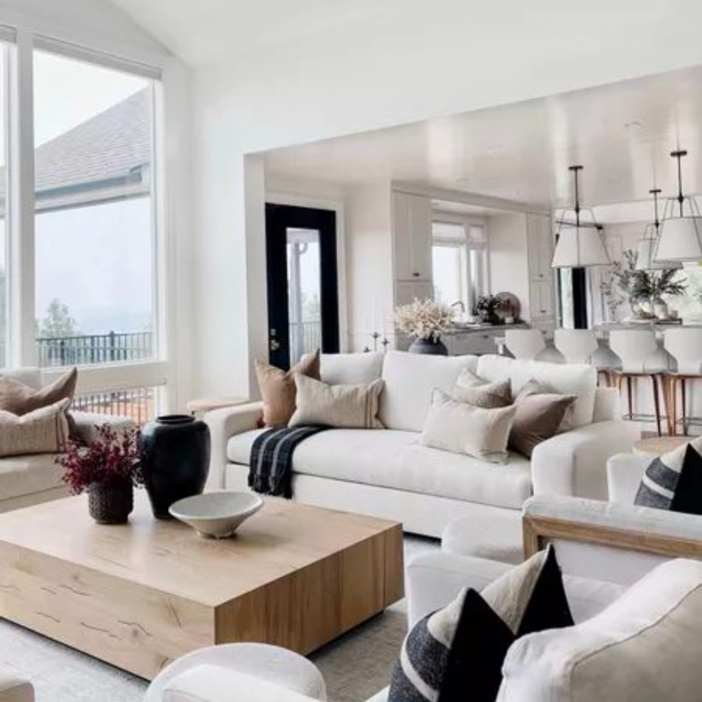 A neutral color palette living room space with an open plan into the kitchen.  The all-white kitchen has an island with four counter stools. 